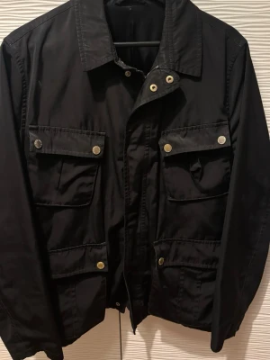 Svart utilityjacka med fickor - Stilren svart utilityjacka med fyra stora fickor framtill och snygga guldfärgade tryckknappar. Klassisk krage och rak passform. Perfekt för dig som gillar en enkel men funktionell look. Materialet känns slitstarkt och jackan har en tidlös design.Revan sitter på insidan.