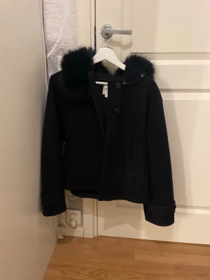 Svart kappa med fuskpäls från Gina Tricot - Fin kappa från Gina tricot med päls på luvan. Storlek 170 så XS/S