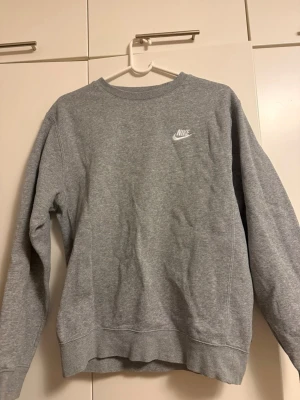 Grå crewneck sweatshirt från Nike - Snygg grå sweatshirt från Nike med klassisk broderad logga på bröstet. Tröjan har rund halsringning, långa ärmar och ribbade muddar. Perfekt för en avslappnad och sportig stil. Tillverkad i mjukt bomullsmaterial.