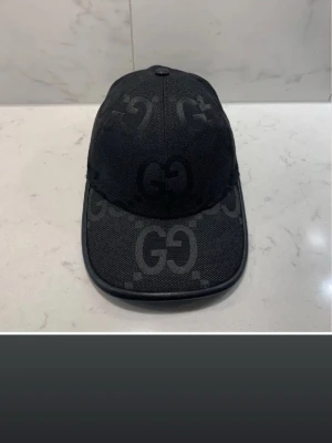 Svart keps från Gucci med GG-mönster - Snygg svart keps från Gucci med klassiskt GG-monogram i mörkgrått över hela kepsen. Kepsen har böjd skärm och är tillverkad i ett slitstarkt tygmaterial. Perfekt för dig som vill ha en stilren och lyxig accessoar.
