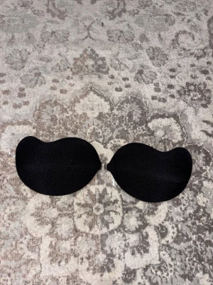 Svart push-up bh från H&M - Säljer en svart push-up bh från H&M med självhäftande insida och transparent spänne framtill. Perfekt för toppar och klänningar med bar rygg eller axlar. Bh:n har en slät och enkel design utan axelband eller ryggband.