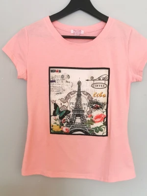 Superfin rosa t-shirt med tryck, storlek S - Snygg rosa t-shirt från Pop Cafe med korta ärmar och ett stort tryck framtill av Eiffeltornet, blommor och fjäril. T-shirten har en klassisk passform, perfekt för dig som gillar Paris-vibbar och coola prints. Har använts 1-2 gånger, är i nyskick med andra ord. Storlek S
