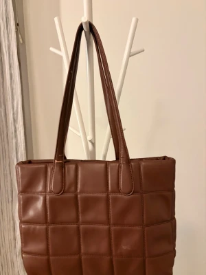 Brun quiltad handväska i läderimitation - Snygg brun handväska med quiltat rutmönster och långa axelband. Väskan är tillverkad i läderimitation och har en stilren, fyrkantig form. Perfekt för dig som gillar trendiga accessoarer med lite extra struktur.