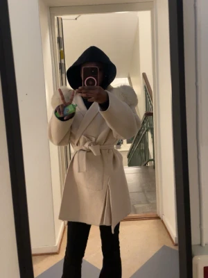 Beige kappa med pälskrage och knytskärp - Snygg beige kappa med bred pälskrage och knytskärp i midjan. Kappan har en klassisk omlott-design och är tillverkad i ett mjukt material som ger en lyxig känsla. Perfekt för att hålla sig varm och stilren under kalla dagar. ÄKTAPÄLS