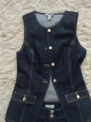Mörkblå jeansväst - Snygg mörkblå jeansväst med markerad midja och dekorativa kontrastsömmar. Västen har fyra ljusa knappar framtill. Ärmlös och tillverkad i denim för en cool och tidlös look.