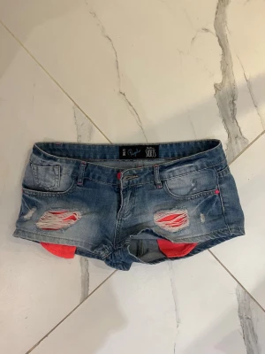 Low waist shorts - Säljer ett par blå jeansshorts från Chillin med slitna detaljer och råa kanter. Shortsen har låg midja, klassiska fem fickor och rosa detaljer som sticker fram vid slitningarna. Perfekta för sommaren och ger en avslappnad look.