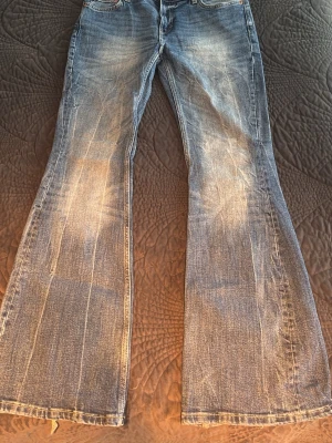 Blå bootcut jeans med slitningar - Ett par blå bootcut jeans med tydliga slitningar och fransade benslut. Jeansen har en klassisk femficksmodell och är tillverkade i denim med en tvättad look. Perfekta för en avslappnad och trendig stil.