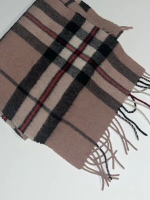 Beige rutig halsduk med fransar - Snygg beige halsduk med klassiskt rutmönster i svart, vitt och rött. Halsduken har mjukt material och avslutas med fransar i ändarna. Perfekt accessoar för att lyfta din outfit och hålla dig varm under kyliga dagar.