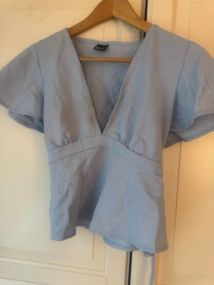 Ljusblå omlottopp med knytband - Söt och somrig topp i en härlig ljusblå färg. Toppen har en smickrande djup V-ringning och korta fladdriga ärmar. Den knyts med ett band i midjan, vilket gör att man kan justera passformen själv.   