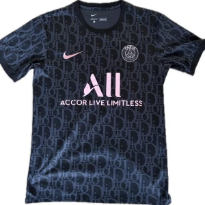 PSG x Dior svart matchtröja - Snygg svart PSG matchtröja från Nike med rosa detaljer och logga. Tröjan har ett mönster med bokstäver över hela tyget, rund halsringning och korta ärmar. Tillverkad i ett lätt och snabbtorkande material, perfekt för fotboll eller träning.