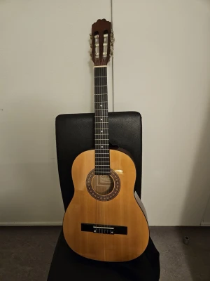 Klassisk gitarr från Cataluña - Säljer en klassisk akustisk gitarr från Cataluña, modell SGN-C-1. Gitarren har en naturlig träfärg med dekorativ rosett runt ljudhålet och sex strängar. Perfekt för dig som vill börja spela eller bara jamma hemma. Helt ny 