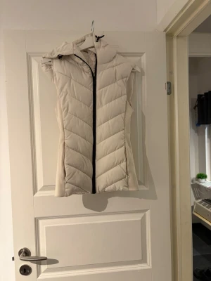Beige dunväst med huva - Stilren beige dunväst med huva och svart dragkedja framtill. Västen har ett quiltat mönster i diagonala ränder och är lätt insvängd för en snygg passform. Perfekt att slänga över en hoodie eller tröja för extra värme. Materialet är mjukt och puffigt. Priset är inte hugget i sten vid snabb affär!