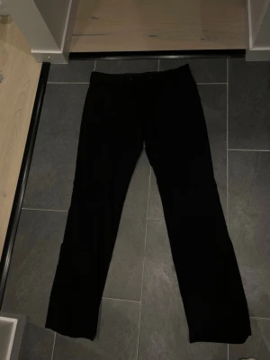 Svarta kostymbyxor med raka ben - Säljer ett par klassiska svarta kostymbyxor med raka ben och diskreta fickor bak. Byxorna har en stilren look och passar perfekt till skjorta eller kavaj. Materialet känns mjukt och följsamt, troligen en blandning av polyester och viskos.