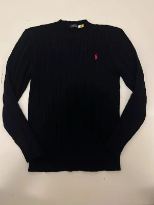 Svart kabelstickad tröja Polo Ralph Lauren - Svart kabelstickad tröja från Polo Ralph Lauren med klassisk röd logga broderad på bröstet. Tröjan har rund halsringning och långa ärmar, perfekt för en stilren och tidlös look.