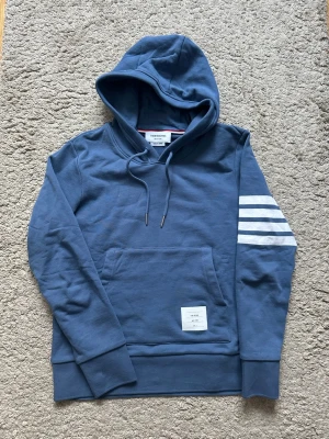 Blå hoodie från Thom Browne - Passar 175cm Fast prisSnygg blå hoodie från Thom Browne med klassiska vita ränder på ena ärmen och en patch vid fickan. Hoodien har dragsko i huvan och en stor magficka. Tillverkad i mjuk bomull och har en avslappnad passform. Perfekt för en chill och stilren look. 