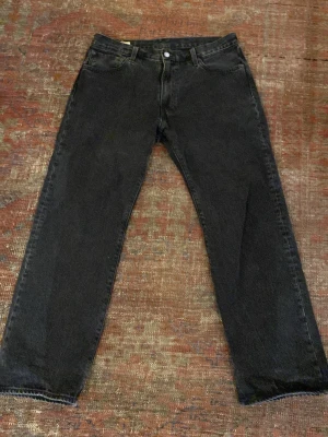 Svarta raka jeans från Levi's - Klassiska svarta jeans från Levi's med rak passform och fem fickor. Jeansen har en enkel design med knappgylf och Levi's ikoniska läderpatch bak i midjan. Tillverkade i slitstark denim som ger en tidlös look. Använda fåtal gånger. Mycket bra skick.