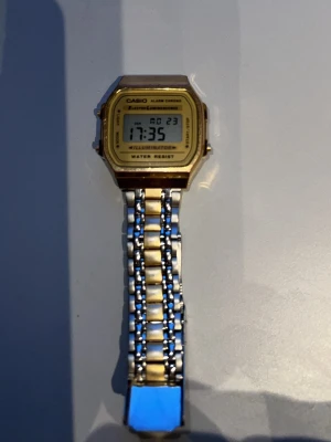 Casio digital guldklocka - Stilren digital klocka från Casio med guldfärgad fyrkantig urtavla och silver- samt guldfärgad länkarmband i metall. Klockan har digital display, alarm och belysning. Perfekt accessoar för dig som gillar retrostil. Helt nytt batteri 