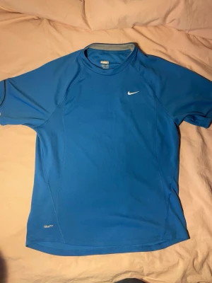 Blå NikeFIT träningströja - Snygg blå träningströja från Nike med korta ärmar och rund hals. Tillverkad i funktionsmaterial som andas, perfekt för gymmet eller löprundan. Diskret Nike-logga på bröstet och NikeFIT-märke nedtill.