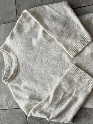 Vit stickad tröja från Gina - Klassisk vit stickad tröja från Gina med rund halsringning och ribbade muddar vid ärmslut och nederkant. Tillverkad i mjukt material som ger en avslappnad och stilren look. Perfekt att matcha med jeans eller kjol.