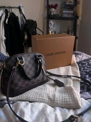 Louis Vuitton Damier Alma BB handväska - Säljer en Louis Vuitton Alma BB handväska i klassiskt brunt Damier-mönster. Väskan har guldfärgade metalldetaljer, lås och dragkedja samt avtagbar axelrem. Tillverkad i canvas med läderdetaljer. Perfekt storlek för det viktigaste och superstylish.