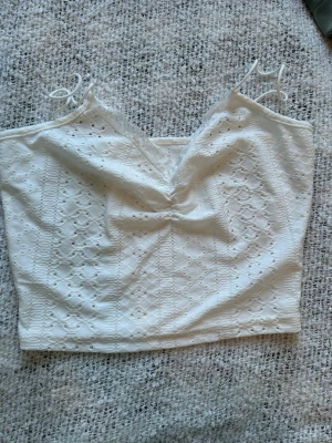 Vit spetsig croppad linnetopp - Söt vit croppad linnetopp med tunna axelband och spetsdetaljer. Toppen har ett broderat hålmönster och en lätt v-ringad form med rynk framtill. Perfekt för sommarens alla tillfällen!