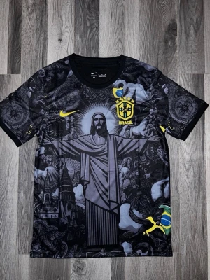 Svart Brasilien Nike fotbollströja - Unik svart fotbollströja från Nike med Brasilien-emblem och gul logga. Framsidan har ett stort motiv av Kristusstatyn och detaljerade mönster i grått. Materialet är lätt och andas, perfekt för träning eller match. Korta ärmar och rund hals.
