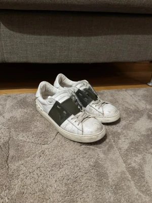 Valentino garvani 40 - Säljer ett par vita sneakers från Valentino Garavani med bred svart rem över vristen och klassisk låg siluett. Skorna har vita snören och detaljer i läder. Perfekta för dig som gillar exklusiv streetstyle och vill sticka ut med en ikonisk design.