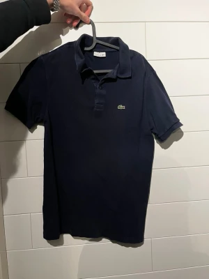 Lacoste pikétröja - Klassisk mörkblå pikétröja från Lacoste med krage och knappar framtill. Tröjan har korta ärmar och den ikoniska gröna krokodilloggan på bröstet. Perfekt för en stilren och avslappnad look.