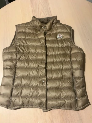 Beige/guld dunväst från Moncler - Dunväst från Moncler med klassisk quiltad design och tryckknappar framtill. Västen har hög krage och ikonisk Moncler-logga på bröstet. Perfekt för lager-på-lager och tillverkad i glansigt material med dunfyllning. Storlek 3 motsvarar M. 