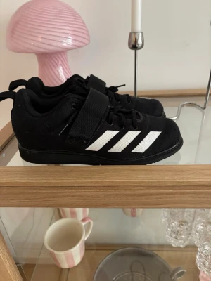 Adidas Powerlift svarta träningsskor - Säljer ett par svarta Adidas Powerlift träningsskor med klassiska vita ränder på sidan. Skorna har kardborreband över snörningen för extra stabilitet och en robust sula som passar perfekt för styrketräning och gym. 