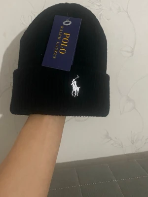 Svart ribbad mössa Polo Ralph Lauren - Snygg svart ribbstickad mössa från Polo Ralph Lauren med klassisk vit broderad logga framtill. Mössan har uppvikt kant och är tillverkad i mjukt material som håller dig varm. Perfekt accessoar för en stilren och enkel look.