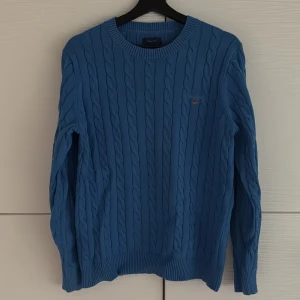 Blå kabelstickad tröja Gant - Snygg blå kabelstickad tröja från Gant med rund halsringning och diskret logga på bröstet. Tröjan har långa ärmar och ribbade muddar vid ärmslut och nederkant. Perfekt för dig som gillar klassisk och stilren soprtig look.