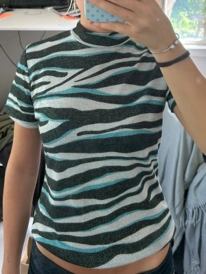 Glittrig zebramönstrad topp - Snygg glittrig topp med zebramönster i svart, silver och turkos. Modellen har korta ärmar och en högre rund halsringning. Perfekt för dig som vill sticka ut med ett coolt mönster och lite extra glitter.
