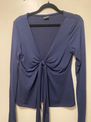 Mörkblå omlottopp med knyt från Gina Tricot - Trendig mörkblå omlottopp med lång ärm och djup v-ringning. Toppen har knyt framtill som ger en snygg och figurnära passform. Perfekt att styla med jeans eller kjol för en cool look.