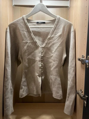 Beige kofta med knappar från Gina Tricot - Stilren beige kofta från Gina Tricot med djup V-ringning och dekorativa knappar framtill. Långärmad och figurnära modell i mjukt material, perfekt för lager-på-lager. Enkel att matcha med olika outfits.