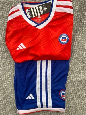 Chile landslagströja Adidas röd/blå för sex åring - Säljer en officiell Chile landslagströja från Adidas i rött och blått med vita detaljer. Tröjan har V-ringning, broderad landslagsemblem och Adidas-logga på bröstet samt klassiska vita ränder på axlar och shorts. Tillverkad i lätt och ventilerande material.