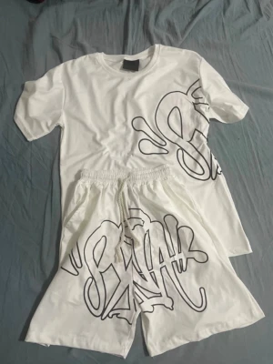Vitt set med graffiti-tryck - Säljer ett vitt set med t-shirt och shorts i mjukt material. Båda plaggen har stora svarta graffiti-inspirerade tryck som ger en cool streetstyle-känsla. T-shirten har rund hals och korta ärmar, shortsen har resår i midjan med dragsko.
