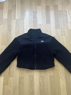 Svart teddyjacka från Alo - Snygg svart teddyjacka från Alo med hög krage och dragkedja framtill. Jackan har en mjuk, fluffig teddystruktur och slät panel upptill med diskret Alo-logga på bröstet. Perfekt för dig som gillar en sportig och trendig look.
