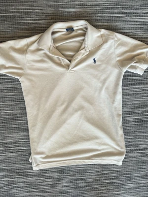 Beige pikétröja från Ralph Lauren - Klassisk beige pikétröja från Ralph Lauren med korta ärmar och den ikoniska blå loggan broderad på bröstet. Tröjan har krage och knappslå, perfekt för en clean och stilren look.