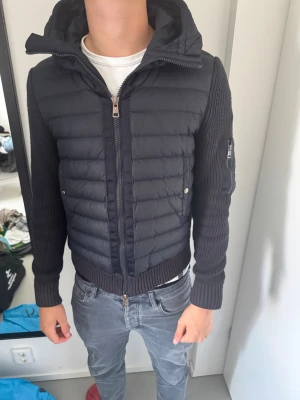 Mörkblå Moncler cardiganjacka - Snygg mörkblå cardiganjacka från Moncler med vadderad front och stickade ärmar. Jackan har huva, dragkedja framtill, fickor med tryckknappar och en extra ficka med dragkedja på ärmen. Moncler-logga på vänster ärm. Perfekt för lager på lager och streetwear. Lite liten på mig som är 187 skulle passa perfekt på dig som är runt 180