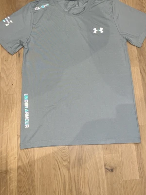 Grå sport-t-shirt från Under Armour - Grå sport-t-shirt från Under Armour med korta ärmar och rund hals. T-shirten har tryckt logga på bröstet och texten UNDER ARMOUR längs sidan. Materialet är lätt och andas, perfekt för träning och aktivitet.