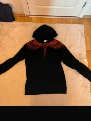 Marcelo burlon  - Säljer en svart hoodie från Marcelo Burlon med ikoniskt rött vingtryck över axlar och bröst. Hoodien har huva med dragsko och ribbade muddar. Perfekt för dig som vill sticka ut med en unik och trendig design.