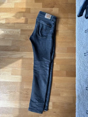 Mörkgrå jeans från Nudie Jeans - Snygga mörkgrå jeans från Nudie Jeans med klassisk femficksdesign och raka ben. Jeansen har en diskret logga på bakfickan och är tillverkade i mjuk bomull med lite stretch för extra komfort.