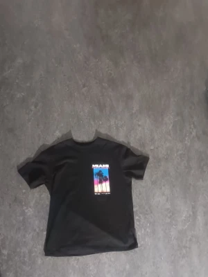 Svart Miami t-shirt med tryck - Cool svart t-shirt med färgglatt Miami-tryck på både fram- och baksidan. Motivet visar palmer mot en solnedgång i blått, rosa och orange. Perfekt för dig som gillar streetstyle och vill sticka ut med en snygg grafisk design.