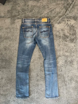 Nudie jeans grim trim  - Snygga blå jeans från Nudie Jeans med klassisk femficksdesign och kontrastsömmar. Jeansen har en ljus tvätt och ikonisk vågdetalj på bakfickorna samt läderpatch med logga i midjan. Perfekta för en avslappnad och trendig look.