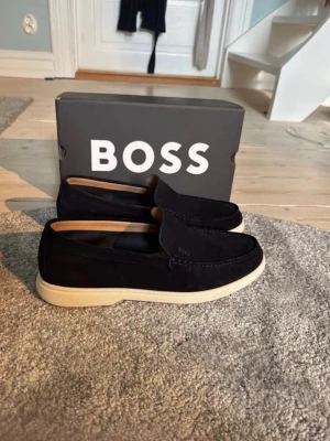 Mörkblå loafers från BOSS - Stilrena mörkblå loafers från BOSS i mocka med vit sula och diskret logga på sidan. Skorna har en klassisk slip-on design och är fodrade med ljust läder invändigt. Perfekta för en clean och avslappnad look.