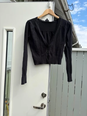 Svart cropped kofta med v-ringning - Svart, tunnstickad kofta med v-ringning och långa ärmar. Modellen är cropped och har ribbade muddar samt knäppning framtill med två knappar. Perfekt för lager-på-lager och enkel att matcha till olika outfits.