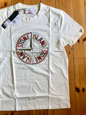 Vit Stone Island x Supreme t-shirt - Vit t-shirt från Stone Island x Supreme med stort broderat färgglatt logomärke på bröstet. Klassisk rund hals och korta ärmar. Perfekt för dig som gillar streetwear och vill sticka ut med en unik design.