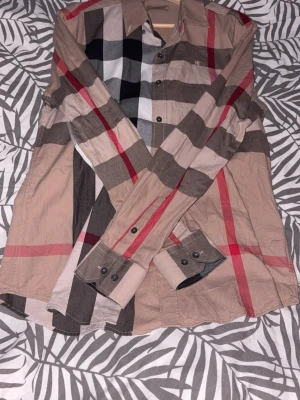 Rutig skjorta från Burberry - Snygg rutig skjorta från Burberry i klassiska färger som beige, svart, vit och röd. Skjortan har långa ärmar, knappar framtill och klassisk krage. Perfekt för dig som vill sticka ut med en ikonisk design och lyxig känsla.(REP) har också bara använts vid ett tillfälle.