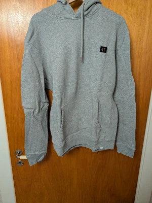 Grå hoodie från Les Deux - Snygg grå hoodie från Les Deux med broderad logga på bröstet. Klassisk känguruficka framtill, justerbar dragsko i huvan och ribbade muddar. Perfekt för en avslappnad och stilren look.